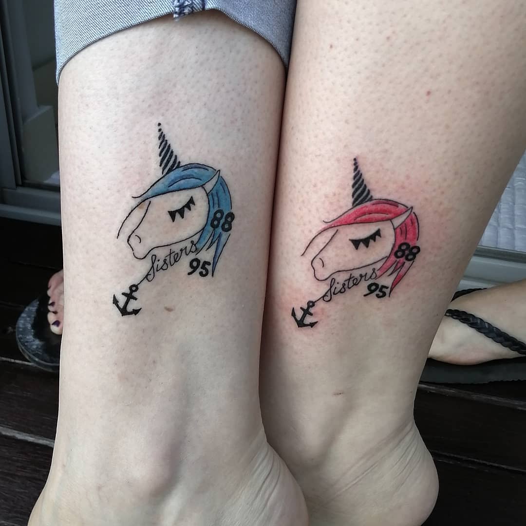 Two ankles with similar unicorn tattoos with blue and pink manes and the word "sisters"